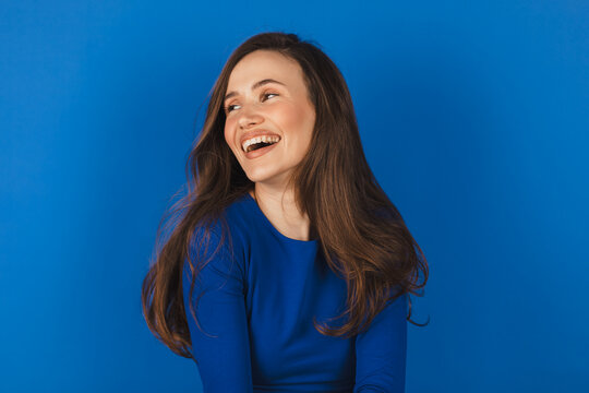 Happy cheerful young woman wearing blue long sleeves top rejoicing at positive news or birthday gift, looking at camera with joyful and charming smile. Brunette student girl isolated blue look at side