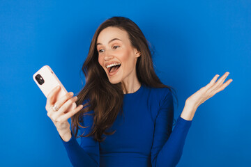 Overjoyed happy excited woman say wow, glad to receive text message informing about salary, rejoices good news, stares at mobile phone, gestures actively from happiness isolated blue background.