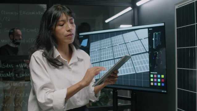 Side pull out footage of female Hispanic engineer entering data about photovoltaic cells into digital tablet looking at solar panel model during workday in renewable energy department