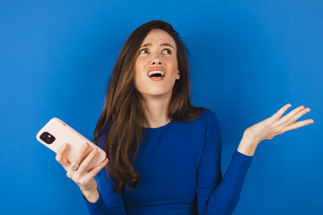 Mad and shock young brunette beautiful woman using mobile phone isolated on blue background....