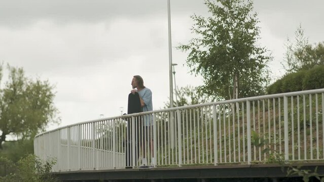 Wide shot of Caucasian adult male wake surfer standing with board on bridge and looking at water on cloudy day