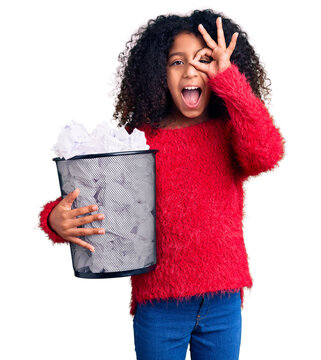 African american child with curly hair holding paper bin full of crumpled papers smiling happy doing ok sign with hand on eye looking through fingers