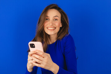 Attractive cheerful amazed girl using device gadget app, sms, post, share social media news isolated blue background. Positive woman holding gadget, texting, messaging. Girl wear blue long sleeves top