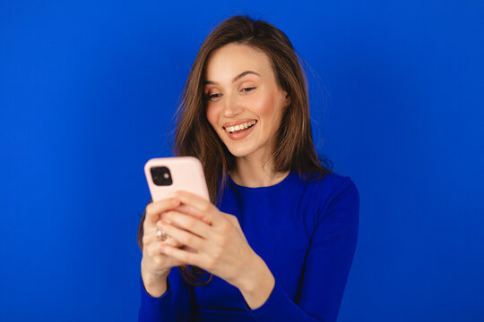 Attractive cheerful amazed girl using device gadget app, sms, post, share social media news isolated blue background. Positive woman holding gadget, texting, messaging. Girl wear blue long sleeves top