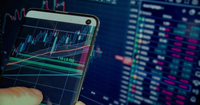 business man checks the chart with financial candles on his smartphone with business news on a computer screen in the background