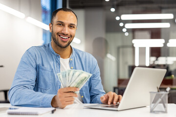 Fototapeta premium Portrait of a smiling young Latin American man sitting at an office desk, working on a laptop, holding cash banknotes in his hand, looking confidently at the camera