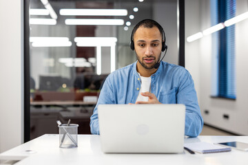 Latin American man in a headset sits in the office in front of a laptop and communicates on a video call - obrazy, fototapety, plakaty