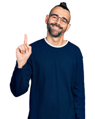 Fototapeta premium Hispanic man with ponytail wearing casual sweater and glasses showing and pointing up with finger number one while smiling confident and happy.