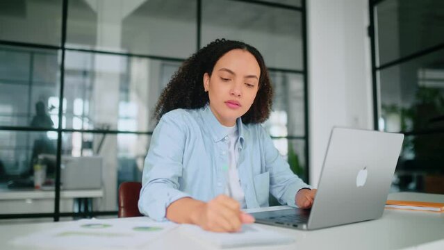 Motivated focused gorgeous brazilian or hispanic curly woman in a pastel blue shirt, employee of a company, sit in modern office at work desk, working on a laptop, concentrated takes notes in notebook