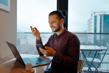 Portrait of smiling handsome man explains business through video chatting working at laptop, holding smartphone in hand, background of panoramic windows.