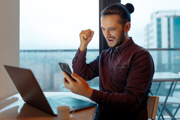 Portrait of handsome man feeling happy, doing winner gesture, video chatting on smartphone with the client, working at laptop, background of panoramic windows.