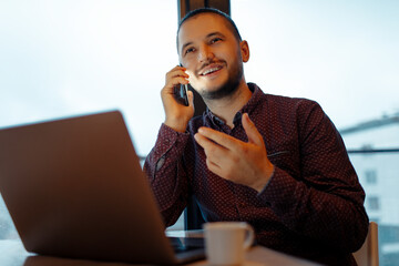 Portrait of smiling handsome man talking on smartphone, explains businesses, working at laptop, background of panoramic windows.