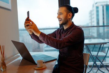 Portrait of happiness handsome man making selfie photo on smartphone, working at laptop, background of panoramic windows.