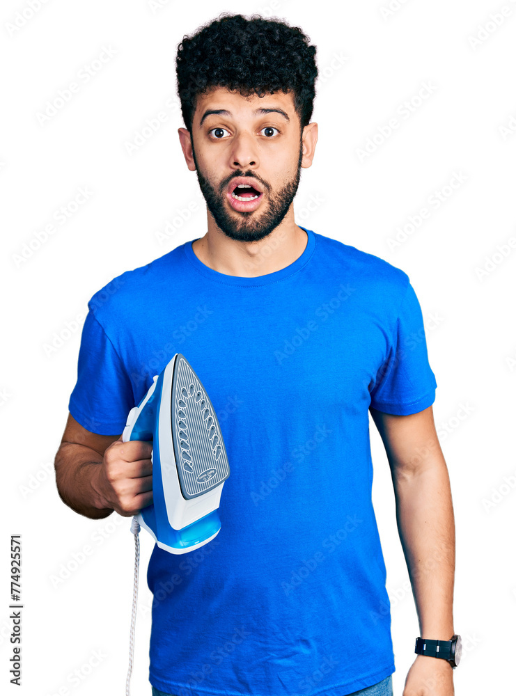 Sticker young arab man with beard holding electric steam iron scared and amazed with open mouth for surprise