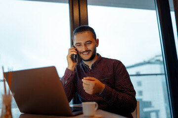 Portrait of handsome smiling man talking on smartphone, working at laptop, background of panoramic windows.