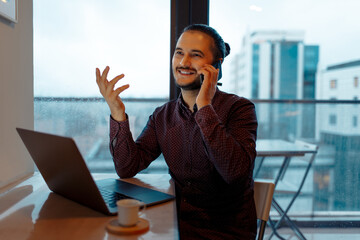 Portrait of young happiness attractive man talking on smartphone, working at laptop, background of panoramic windows.