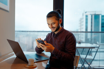 Portrait of smiling handsome man with hair bun working at laptop, using smartphone, background of panoramic windows.