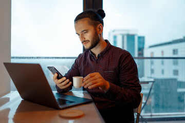 Portrait of confident handsome man working at laptop, looking in smartphone, holding the espresso cup of coffee, background of panoramic windows.