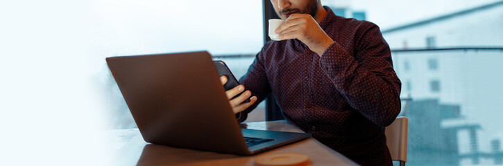 Man working at laptop, drinking the coffee, holding smartphone in hand. Panoramic portrait.
