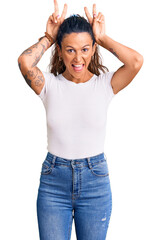 Young hispanic woman with tattoo wearing casual white tshirt posing funny and crazy with fingers on head as bunny ears, smiling cheerful