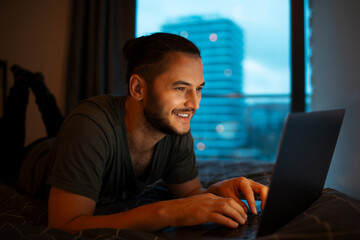 Portrait of young smiling man working at laptop while lying on the bed close to evening time.
