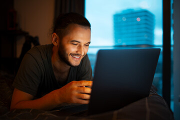 Portrait of young smiling man working at laptop while lying on the bed close to evening time.
