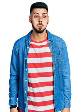 Young hispanic man with beard wearing casual denim jacket puffing cheeks with funny face. mouth inflated with air, crazy expression.