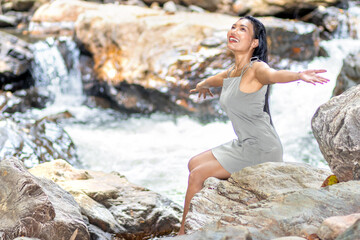 A young woman with outstretched arms sits on boulders in a water cascade