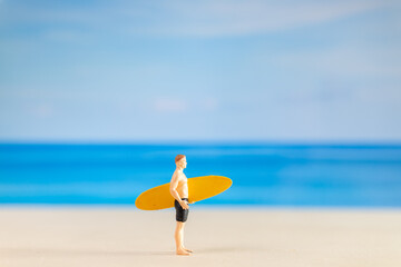 Miniature people man in a swimsuit, and holding a yellow surfboard on the beach