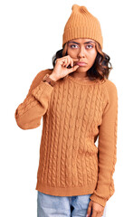 Young beautiful mixed race woman wearing wool sweater and winter hat mouth and lips shut as zip...