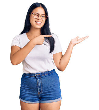 Young beautiful asian girl wearing casual clothes and glasses amazed and smiling to the camera while presenting with hand and pointing with finger.