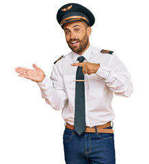 Handsome man with beard wearing airplane pilot uniform amazed and smiling to the camera while presenting with hand and pointing with finger.