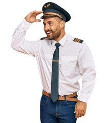 Handsome man with beard wearing airplane pilot uniform very happy and smiling looking far away with hand over head. searching concept.