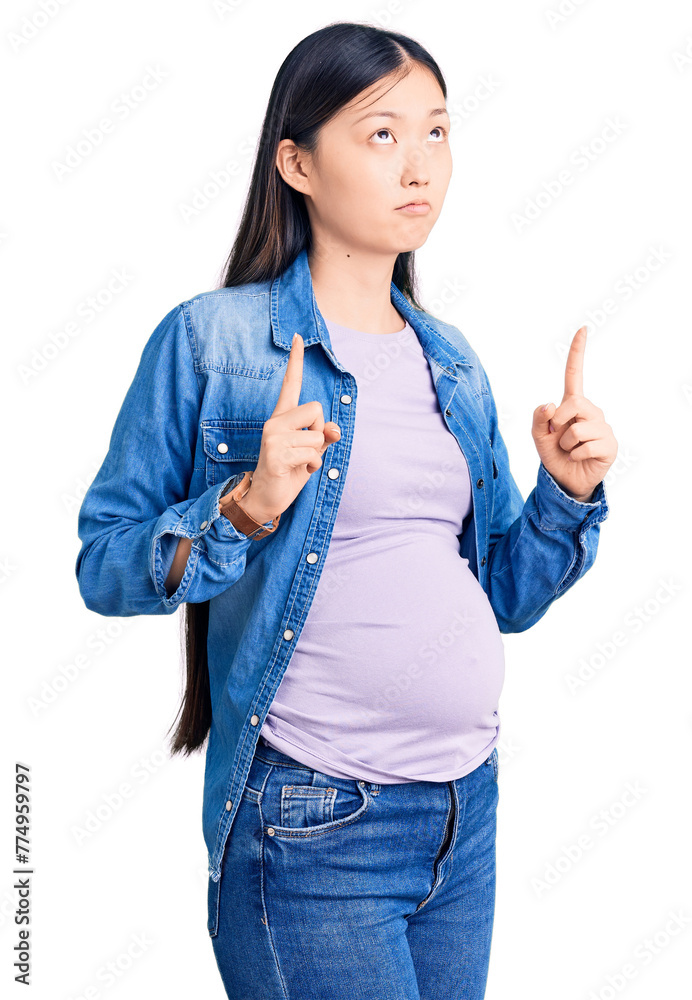 Sticker young beautiful chinese woman pregnant expecting baby pointing up looking sad and upset, indicating 