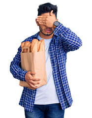 Handsome latin american young man holding paper bag with bread covering eyes with hand, looking...