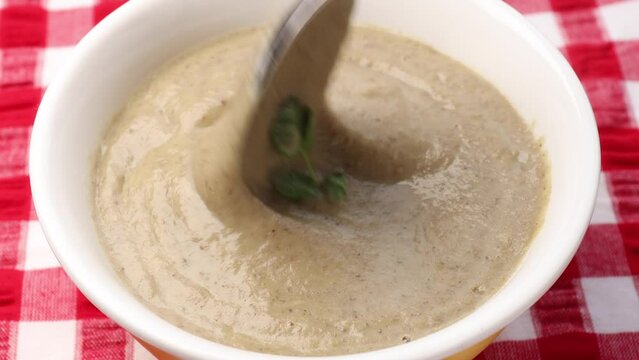 Creamy mushroom soup in a bowl, topped with fresh coriander leaves. Stir the soup with a tablespoon and garnish with slices of saut&eacute;ed mushrooms