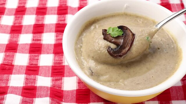 Creamy mushroom soup in a bowl, enhanced with fresh coriander leaves. Taste the soup using a tablespoon, and garnish with slices of sautéed mushrooms.