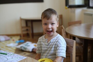 Happy Caucasian boy kid sitting by wooden desk and laughing at a Kindergarten, vertical shot
