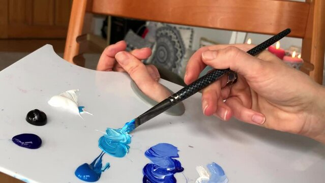 Mixing acrylic paint on white palette. Close up on hands of a female artist mixing blue, white and black acrylic paint with a brush on white palette. High quality 2K footage.