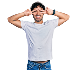 Hispanic young man with beard wearing casual white t shirt doing peace symbol with fingers over face, smiling cheerful showing victory