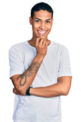 Young hispanic man wearing casual white t shirt looking confident at the camera smiling with crossed arms and hand raised on chin. thinking positive.