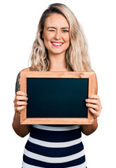 Young blonde woman holding blackboard winking looking at the camera with sexy expression, cheerful...
