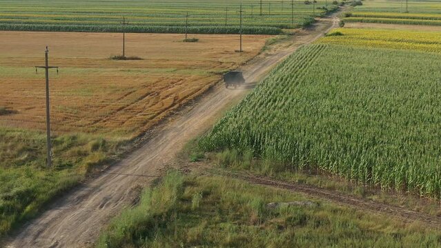 Aerial shot of tractor with trailer driving along the countryside dirt road through cultivated fields in summer afternoon, drone pov