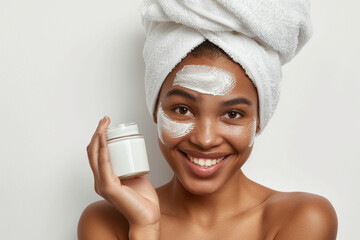 Skincare, cheerful young African American woman smiling, applying moisturizing cream on face, lotion or mask for skin lifting and anti-aging, wears wrapped towel on head, with plastic jar in hand. 