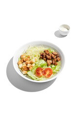 Fresh Caesar salad with crispy croutons and grilled chicken pieces in a white bowl, isolated on a white background with dressing on the side