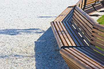 New bench with wooden slats and metal structure in a public park on white gravel pavement - obrazy, fototapety, plakaty