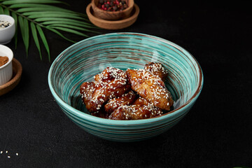 Roasted chicken wings in teriyaki sauce with sesame on ceramic bowl. Buffalo wings in asian style on black concrete background. Aesthetic composition with fried chicken wings, chopsticks and leaf.