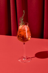 Alcohol cocktail with aperol and thyme on red background with shadow. Aperol spritz cocktail on coloured background in trendy style. Contemporary concept with alcohol beverage. Bartender cocktail.
