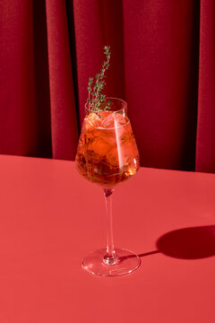 Alcohol cocktail with aperol and thyme on red background with shadow. Aperol spritz cocktail on coloured background in trendy style. Contemporary concept with alcohol beverage. Bartender cocktail.