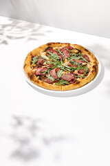 Italian pizza with roast beef, arugula and spicy sauce on light background with shadows. Meat pizza with sliced roast beef on white table. Italian food in trendy menu restaurant